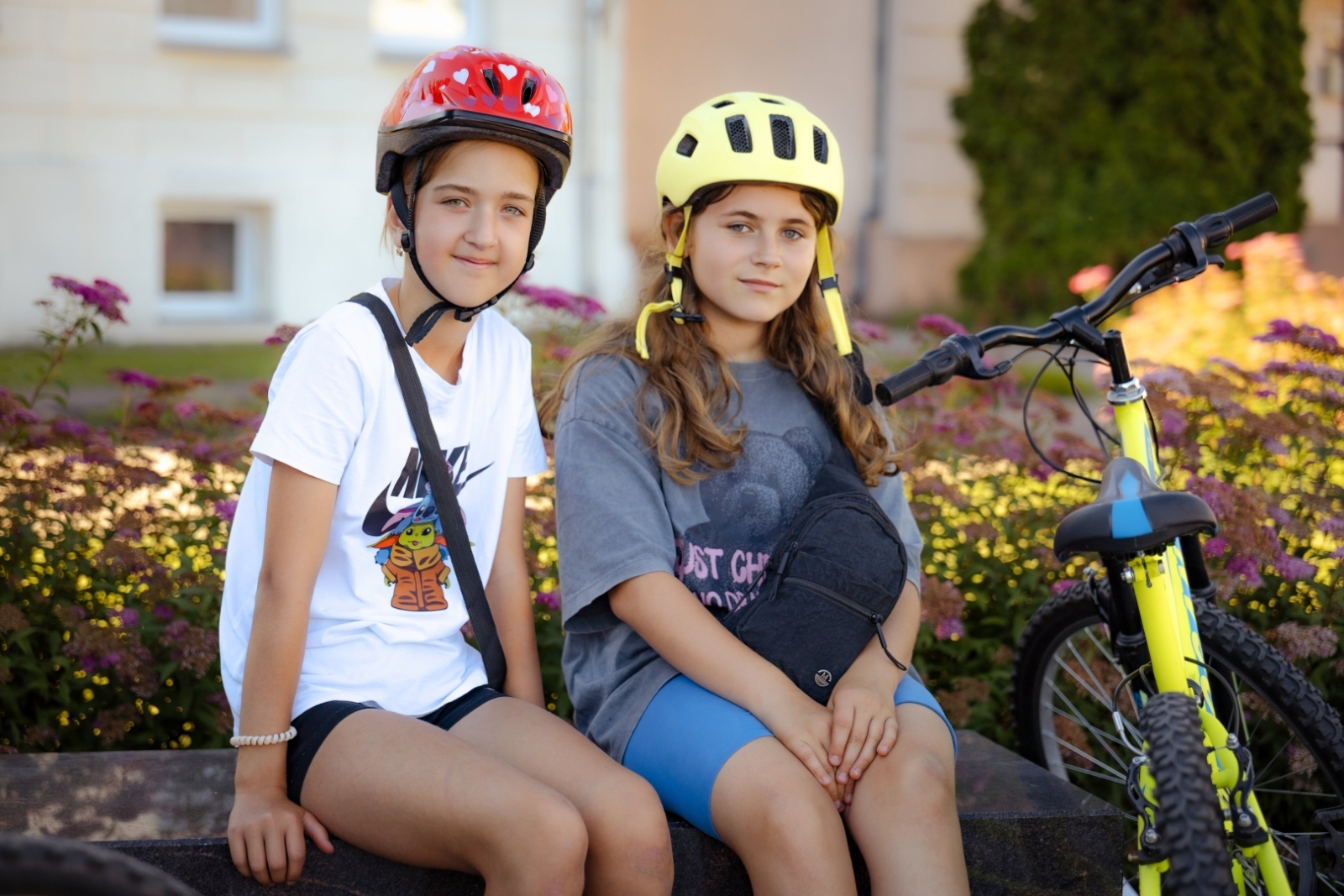 Divas meitenes velosipēdu ķiverēs sēž blakus velosipēdam