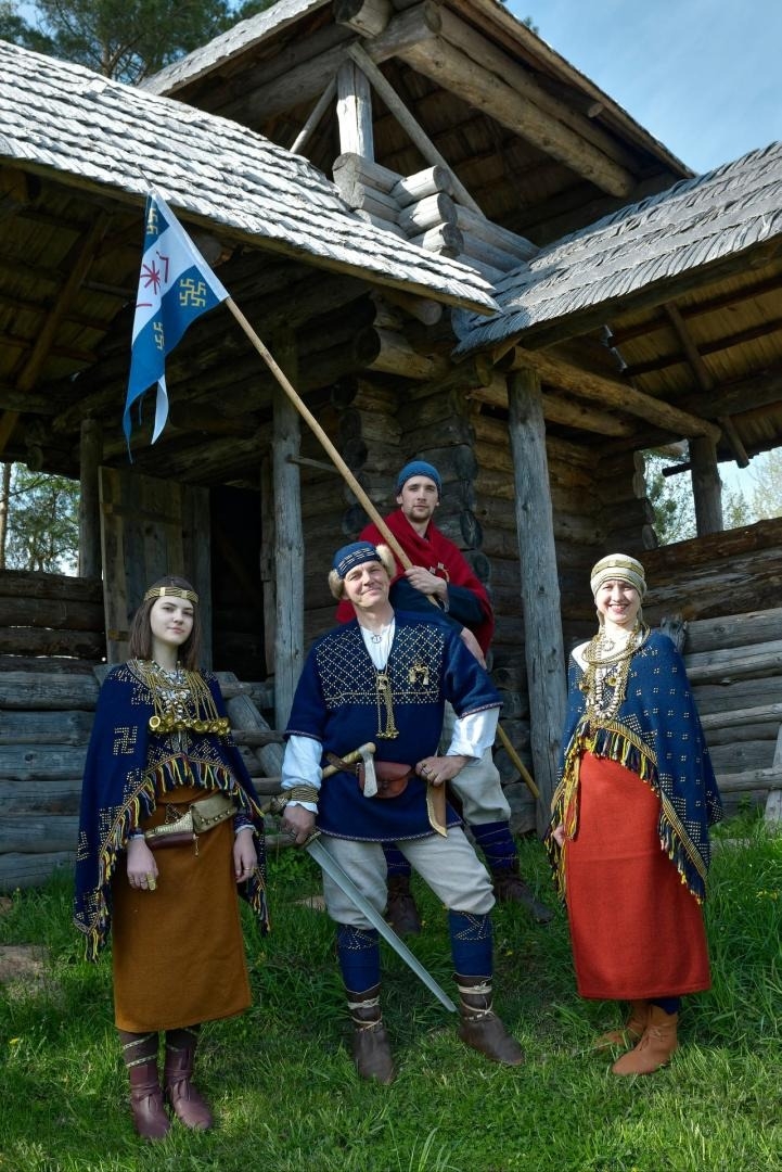 Cilvēki vēsturiskos tērpos pie koka ēkas